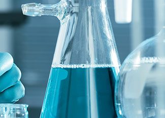 A close-up of a laboratory flask filled with blue liquid, with a gloved hand holding a small container nearby. Laboratory glassware and blurred background suggest a science or research setting.