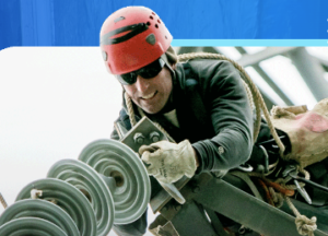 A worker wearing a red helmet, sunglasses, gloves, and safety gear handles electrical insulator discs while working on power lines.