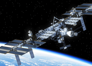 The International Space Station orbits above Earth, with solar panels extended and the blue curve of the planet visible below against the blackness of space.