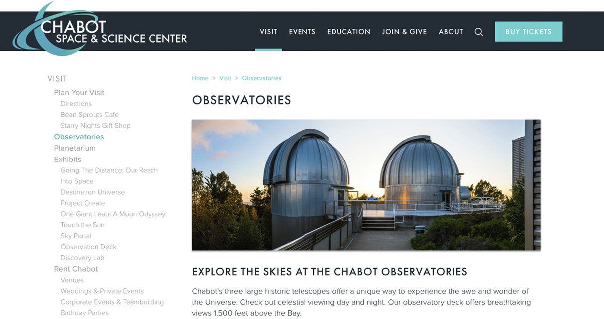 Two silver domed observatories sit side by side on a rooftop platform at sunset, surrounded by trees. The sky is clear with warm, golden light shining on the structures.