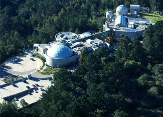 Aerial view of a modern science museum complex with several domed buildings surrounded by dense green trees and a parking lot with cars nearby.