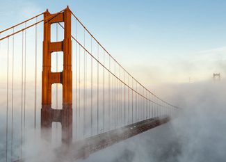 The Golden Gate Bridge towers rise above a thick layer of fog, with the bridge’s cables stretching into the mist under a clear sky.