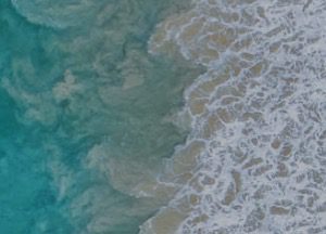 Aerial view of ocean waves crashing onto the shore, with frothy white surf blending into turquoise and sandy-colored water.