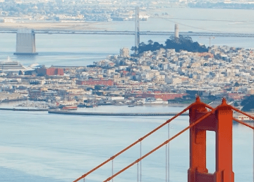 A close-up of the Golden Gate Bridge’s red cables in the foreground, with a cityscape, marina, and distant bridges over the water in the background.