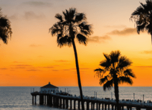 A pier extends into the ocean at sunset, with a small building at the end. Silhouetted palm trees frame the scene against an orange and yellow sky.
