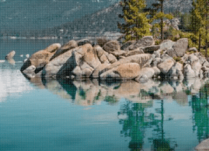 A cluster of large rocks sits at the edge of a clear, turquoise lake, surrounded by pine trees. The calm water reflects the rocks and trees, with mountains visible in the background.