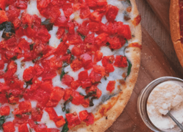 A close-up of a pizza with melted cheese, chopped tomatoes, and herbs on a wooden board next to a small glass jar of grated parmesan cheese.