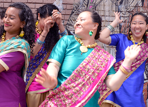 Four women dressed in colorful traditional clothing and jewelry are dancing joyfully outdoors, smiling and raising their arms with a brick wall and mural in the background.