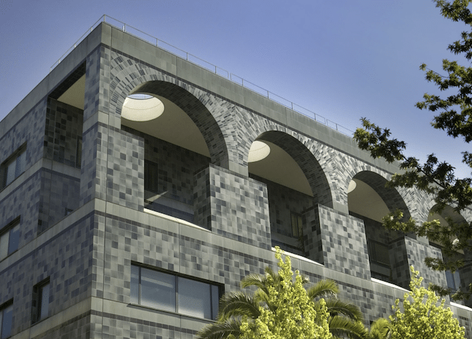 A modern building with a gray tiled facade featuring large arched openings on the upper floor. Palm trees and leafy branches are visible in the foreground against a clear blue sky.