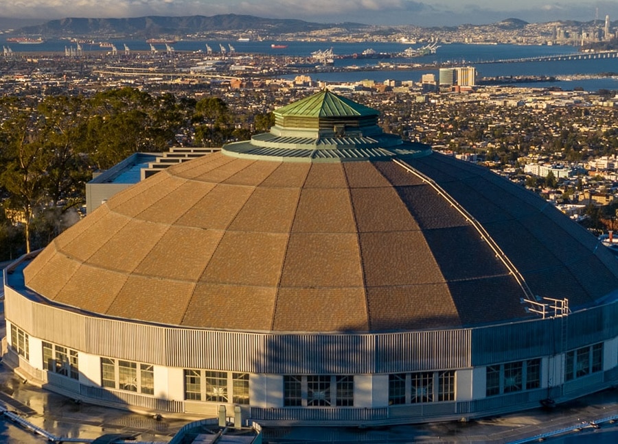A large circular building with a brown dome roof and green central skylight sits on a hillside, overlooking a city, distant bay, ships, and mountains under a partly cloudy sky.