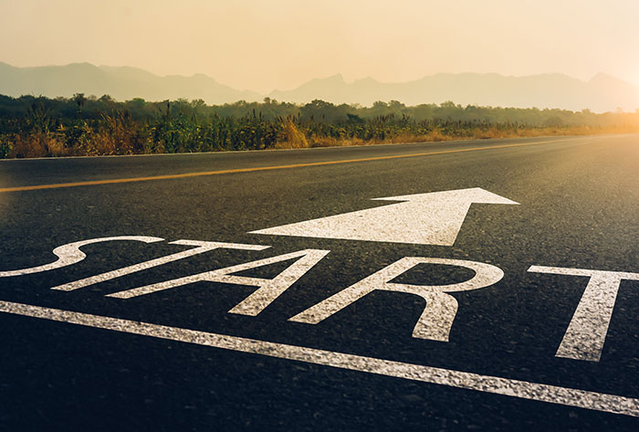 A road with the word START and a large white arrow painted on it, pointing forward. The sun is rising or setting, and mountains and trees are visible in the distance.