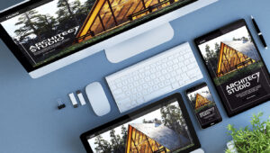 A computer, tablet, smartphone, and laptop display a website for Architect Studio with a triangular wooden house; also shown are a keyboard, mouse, USB drives, headphones, and a small potted plant on a blue desk.