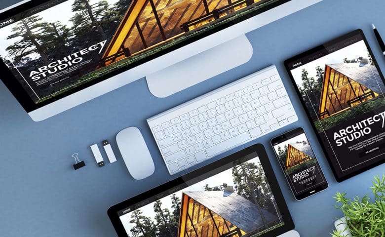 A desktop computer, laptop, tablet, and smartphone display the same Architect Studio website. The devices are arranged neatly on a blue surface, surrounded by a keyboard, mouse, USB drives, and a small potted plant.