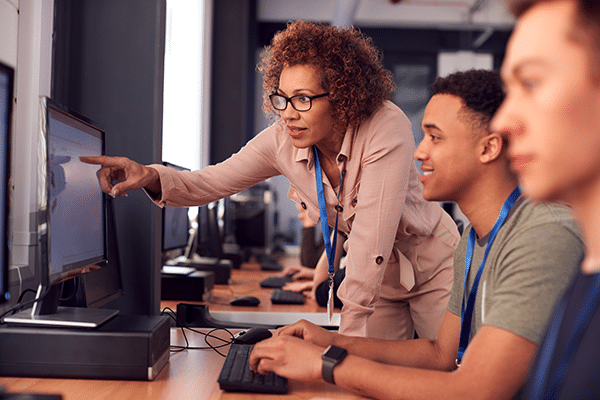 A teacher is standing and pointing at a computer screen, helping two students who are seated and working at computers in a classroom or computer lab setting.