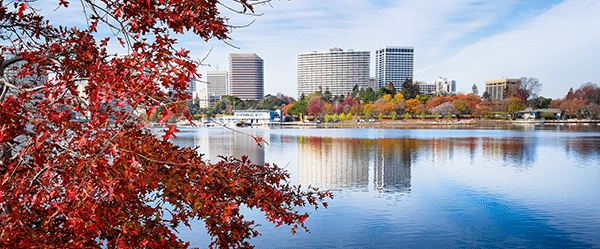 Downtown buildings and colorful autumn trees reflected in a calm lake, with branches of red leaves in the foreground under a partly cloudy sky.