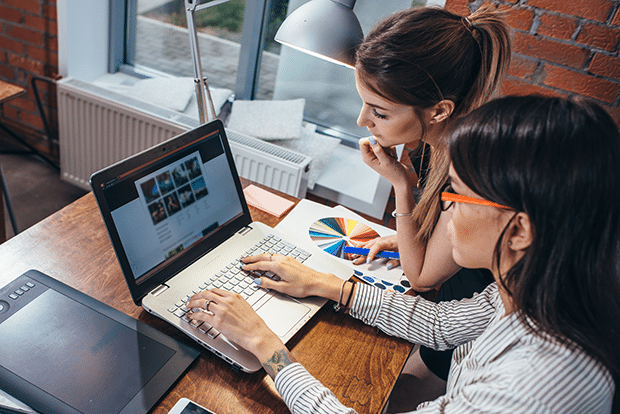 Two women sit at a desk, looking at a laptop screen with images. One woman points at the screen while the other holds a color swatch. A graphic tablet and smartphone are on the desk in a modern office setting.