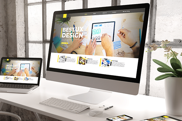 A computer monitor and a laptop on a white desk display a website titled Best UX Design. The workspace includes a plant, smartphone, and office supplies, with sunlight streaming through large windows.