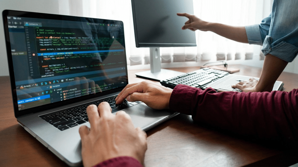 Two people work at a desk; one types code on a laptop, while the other points at a desktop monitor. Both screens display programming text, suggesting collaboration on a software project.