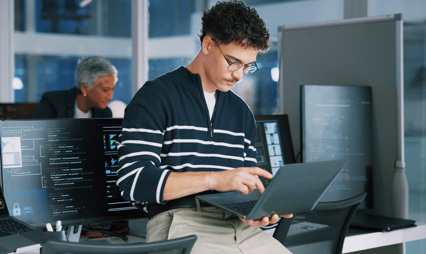 A man with curly hair and glasses is sitting on a desk, working on a laptop. Behind him are computer monitors displaying code, and another person is working in the background in a modern office setting.