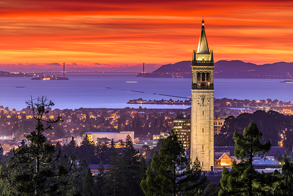 The Campanile tower at UC Berkeley is illuminated at dusk, with city lights, the San Francisco Bay, and the Bay Bridge visible under a vibrant orange and red sunset sky. Trees frame the foreground.