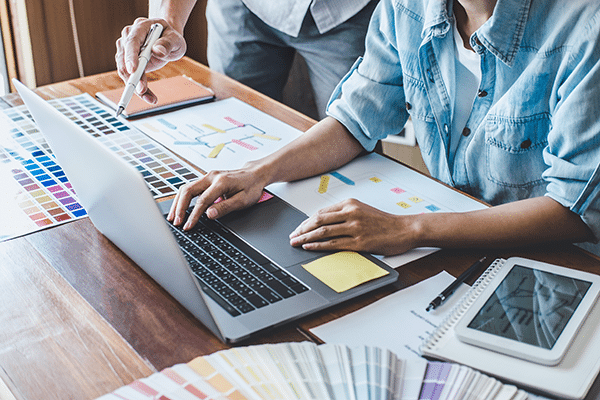 Two people working at a desk with a laptop, color swatches, design sketches, and a notebook. One person is typing on the laptop while the other points at the color samples, suggesting a collaborative creative project.