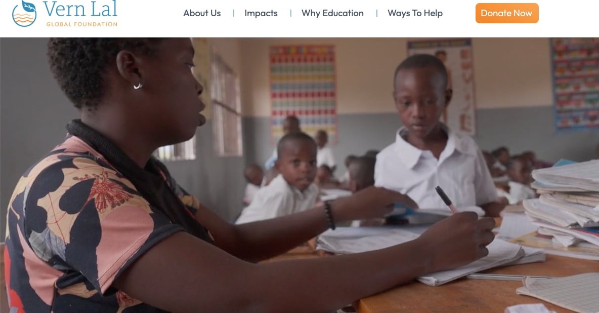 A teacher sits at a desk helping young students in a classroom. The students wear white uniforms and are engaged in their schoolwork. Educational posters are visible on the classroom walls.