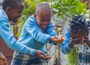 Three children in blue sweaters laugh and play as they gather around an outdoor faucet, catching and drinking water with their hands.