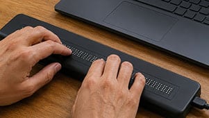 Hands using a refreshable braille display connected to a laptop on a wooden desk, illustrating the importance of tools like these during a WordPress accessibility audit.