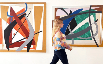 A woman in a colorful sweater walks past two large, abstract paintings with bold shapes and vibrant colors displayed on a white wall.