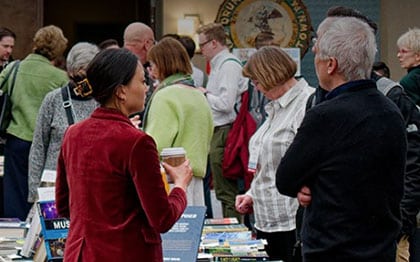 People are browsing tables with books at a busy indoor event. Some are talking, others are looking at the books. One person in the foreground holds a coffee cup. The atmosphere appears lively and social.